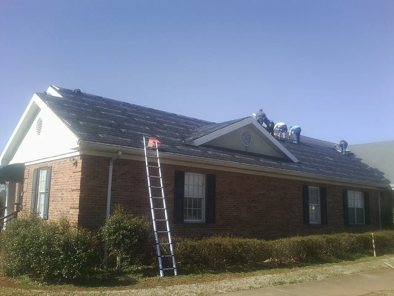 Crew tearing shingles off a brick ranch roof with a ladder set — Metal Roof Repair work by our crew in Great Cacapon