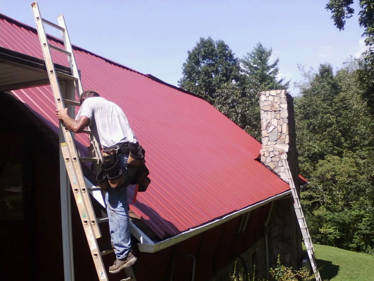 Roofer on a ladder beside a red metal roof and stone chimney — Soffit & Fascia Repair by Roofing Brothers in Great Cacapon